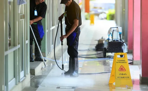 Two workers in black uniforms and boots pressure wash a tiled walkway using a high-pressure water sprayer beside a yellow Cleaning in Progress floor sign.