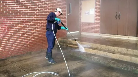 A worker in a blue uniform, cap, and green gloves uses a pressure washer to clean concrete steps outside a red brick building, helping homeowners pressure wash your house and maintain clean, attractive exterior surfaces.