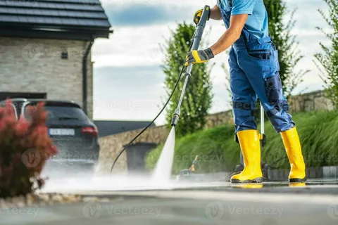 A worker in blue overalls and bright yellow boots uses a high-pressure washer to pressure wash a driveway in front of a modern house and parked car.